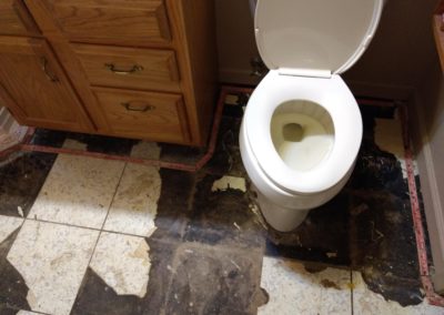 Stained toilet and bathroom floor with carpet tack strips and broken tiles