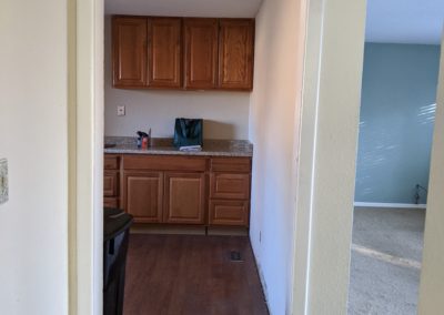 Kitchen cabinets and vinyl floor on left and living room with stained carpet on right with wall and doorway between them