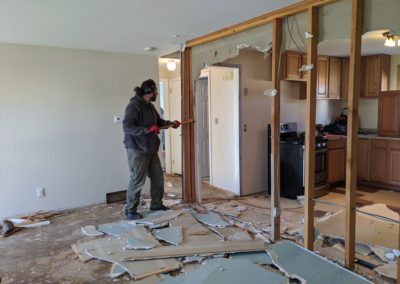 Man uses crowbar to remove framing in corner of demolished wall with some studs removed and drywall debris on subfloor