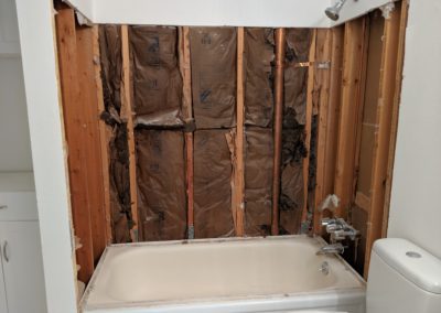 Shower walls stripped to studs and insulation above bathtub with scars from glass door tracks