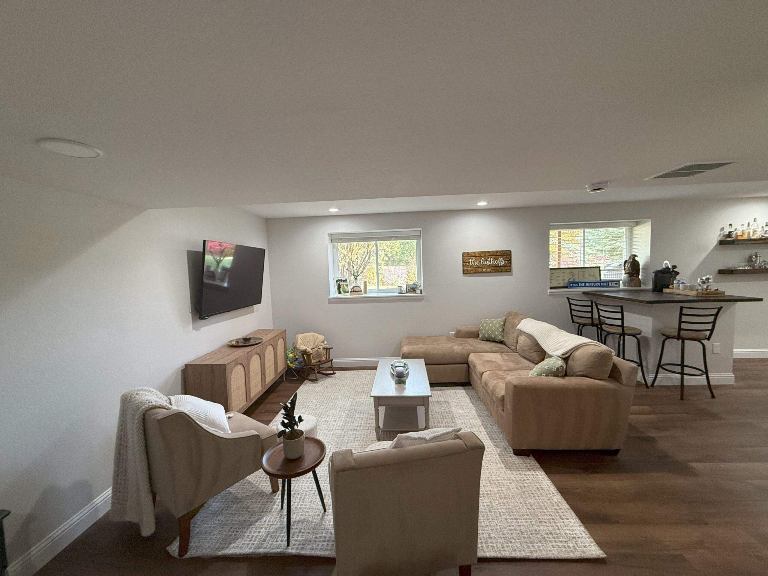 Finished basement sitting area with sectional couch, mounted TV, bar seating, and large windows for natural light.