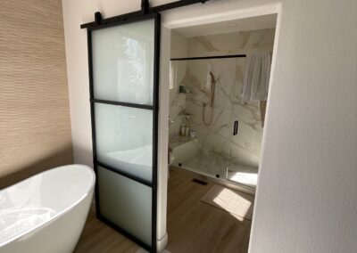Spa-like bathroom with freestanding tub, modern black-framed sliding barn door, and marble-style walk-in shower with brass fixtures.