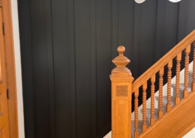 Stairway entry with new board and batten wall painted dark green-black, next to original wood staircase and trim.