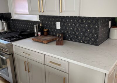 Black geometric tile backsplash with gold accents in a modern kitchen featuring white cabinets and gold hardware.