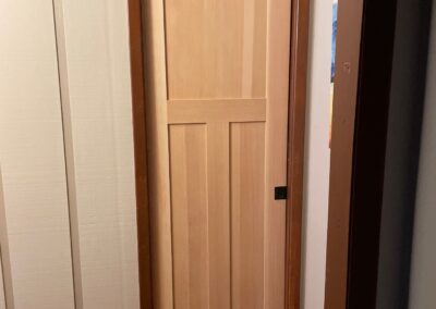 New natural wood 3-panel pocket door installed in a hallway with white vertical paneling and brown trim.