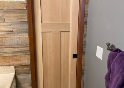 Freshly installed wood pocket door in a bathroom with gray walls, wood accent paneling, and a countertop sink nearby.