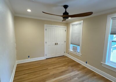 A wide-angle view of a beige bedroom with textured walls, white trim, a ceiling fan, and newly installed wood floors.