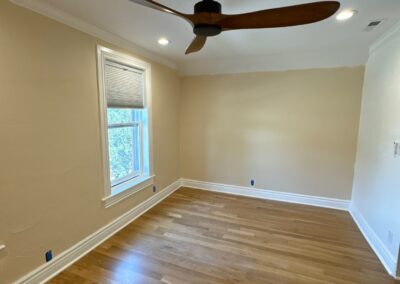 View of an empty room with natural light, beige walls, two windows, and warm wood floors, taken before wall paneling and paint.
