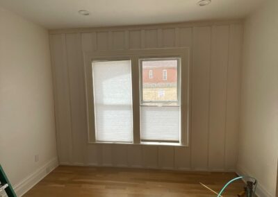 A work-in-progress shot of a room with wood floors, window trim, and visible electrical cords, showing natural light across unfinished walls.