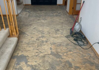 Entryway with old flooring removed, exposing marked-up subfloor and original staircase in background.