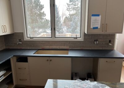 Gray subway tile backsplash being installed above black counters in a kitchen with winter views outside.