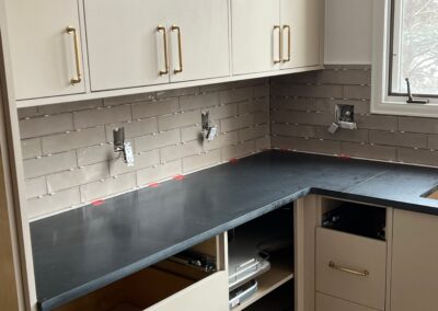 Close-up of corner kitchen cabinets with backsplash tile mid-installation and black countertops below.
