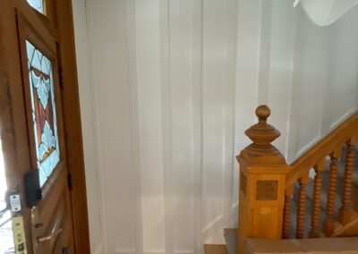 White board and batten paneling installed on stairway wall next to original wood staircase and stained glass front door.