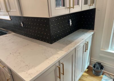Black and gold tile backsplash partially installed along white kitchen cabinets and white marbled countertops.