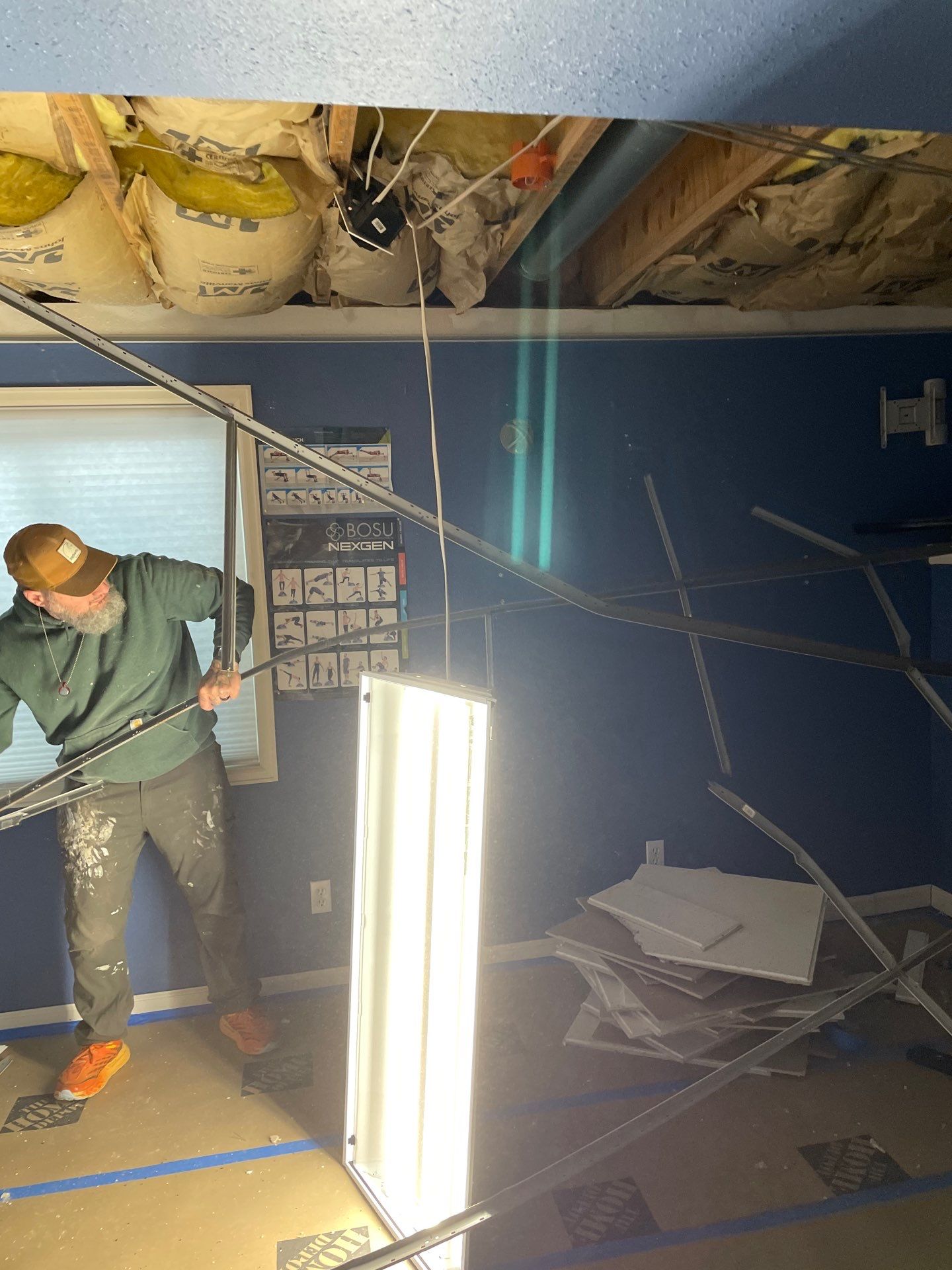 Demo Contractor dismantling a dropped ceiling in a blue room with exposed insulation and hanging fluorescent light.