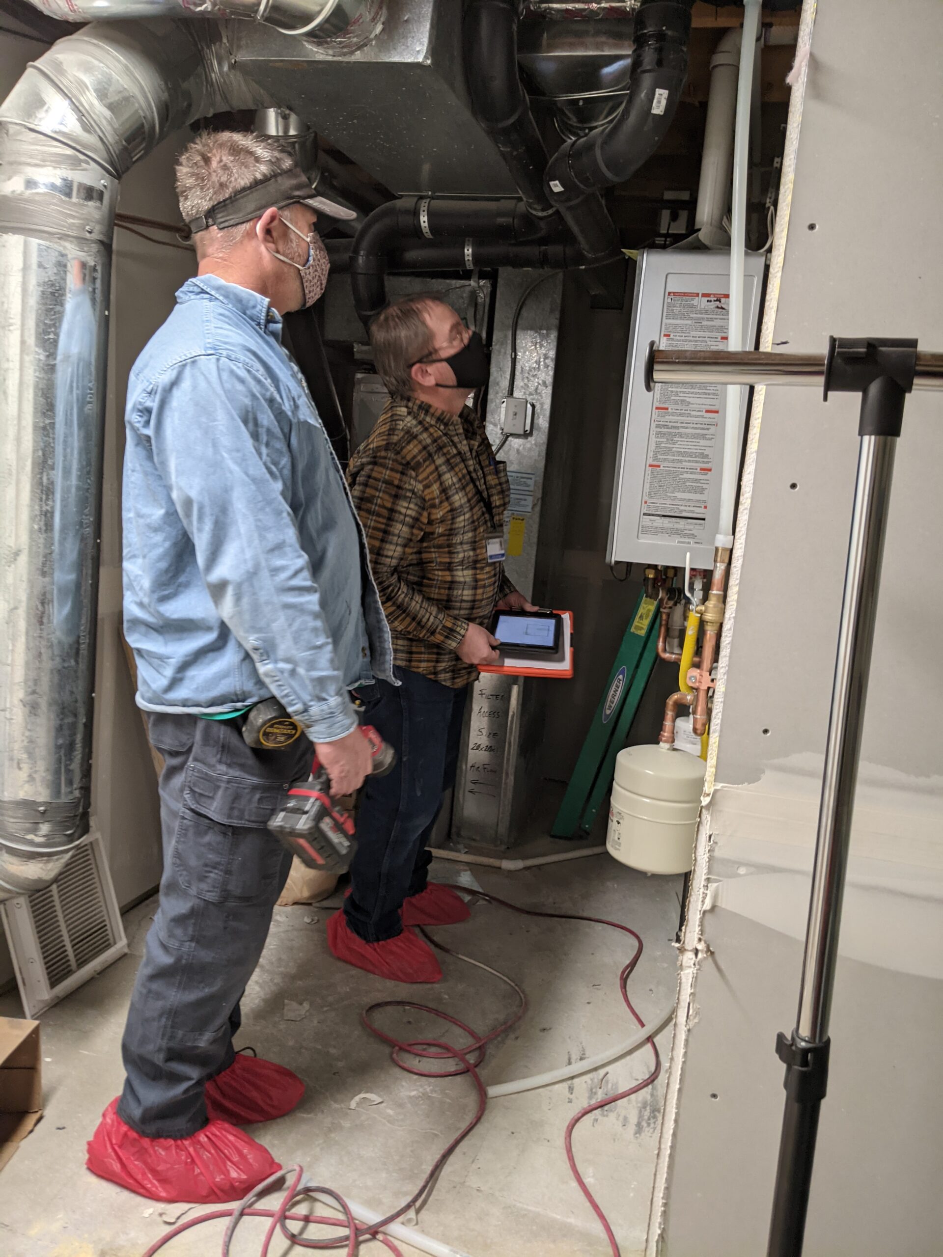 Plumber&Inspector Two men in masks and red shoe covers inspect HVAC and plumbing systems in a utility room.