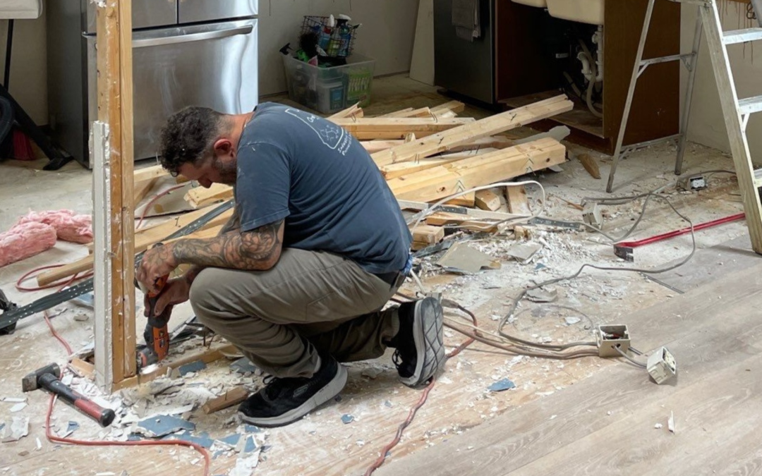Contractor kneeling while dismantling a wood-framed kitchen wall surrounded by debris, tools, and exposed outlets.
