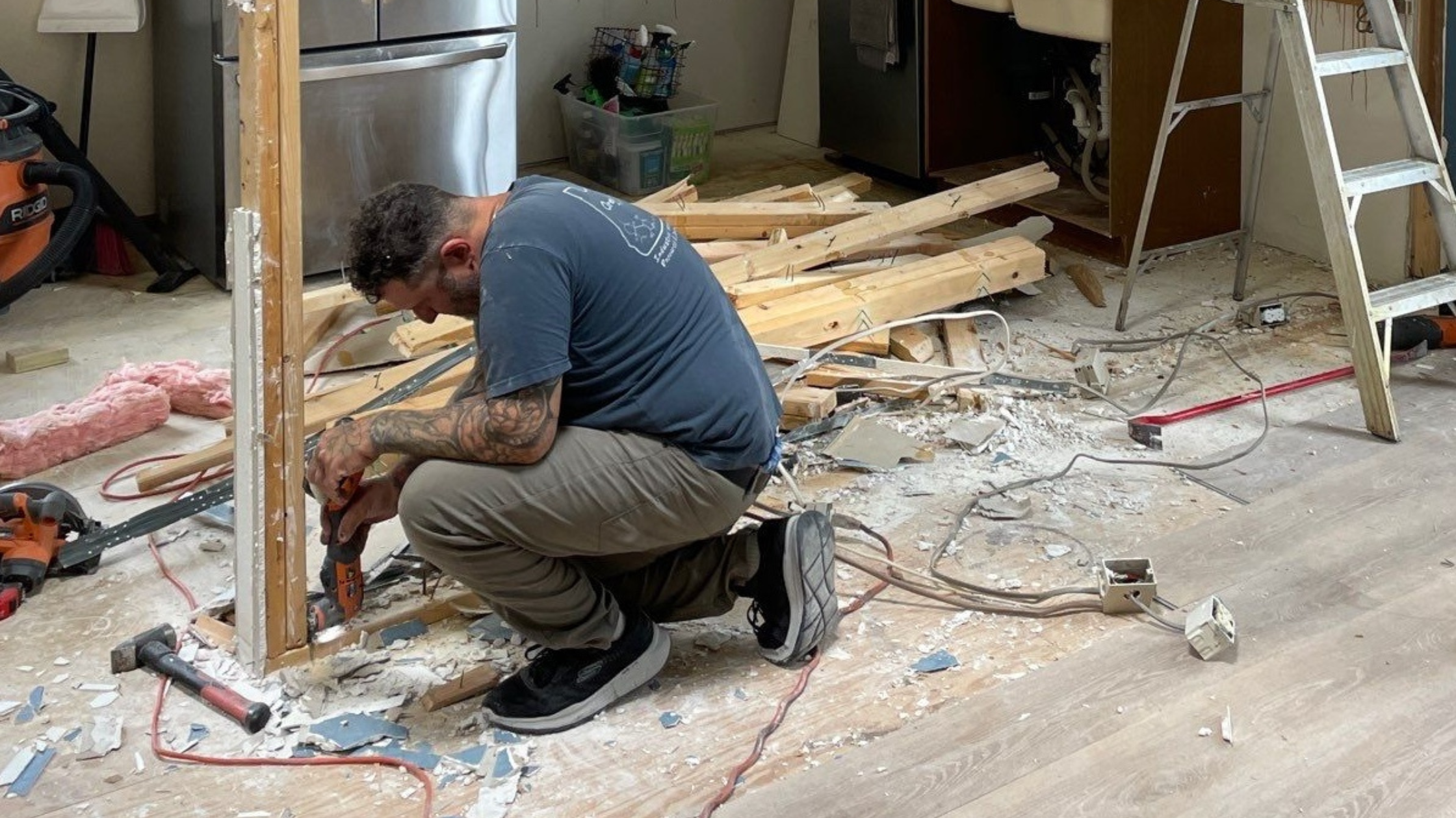 Contractor kneeling while dismantling a wood-framed kitchen wall surrounded by debris, tools, and exposed outlets.