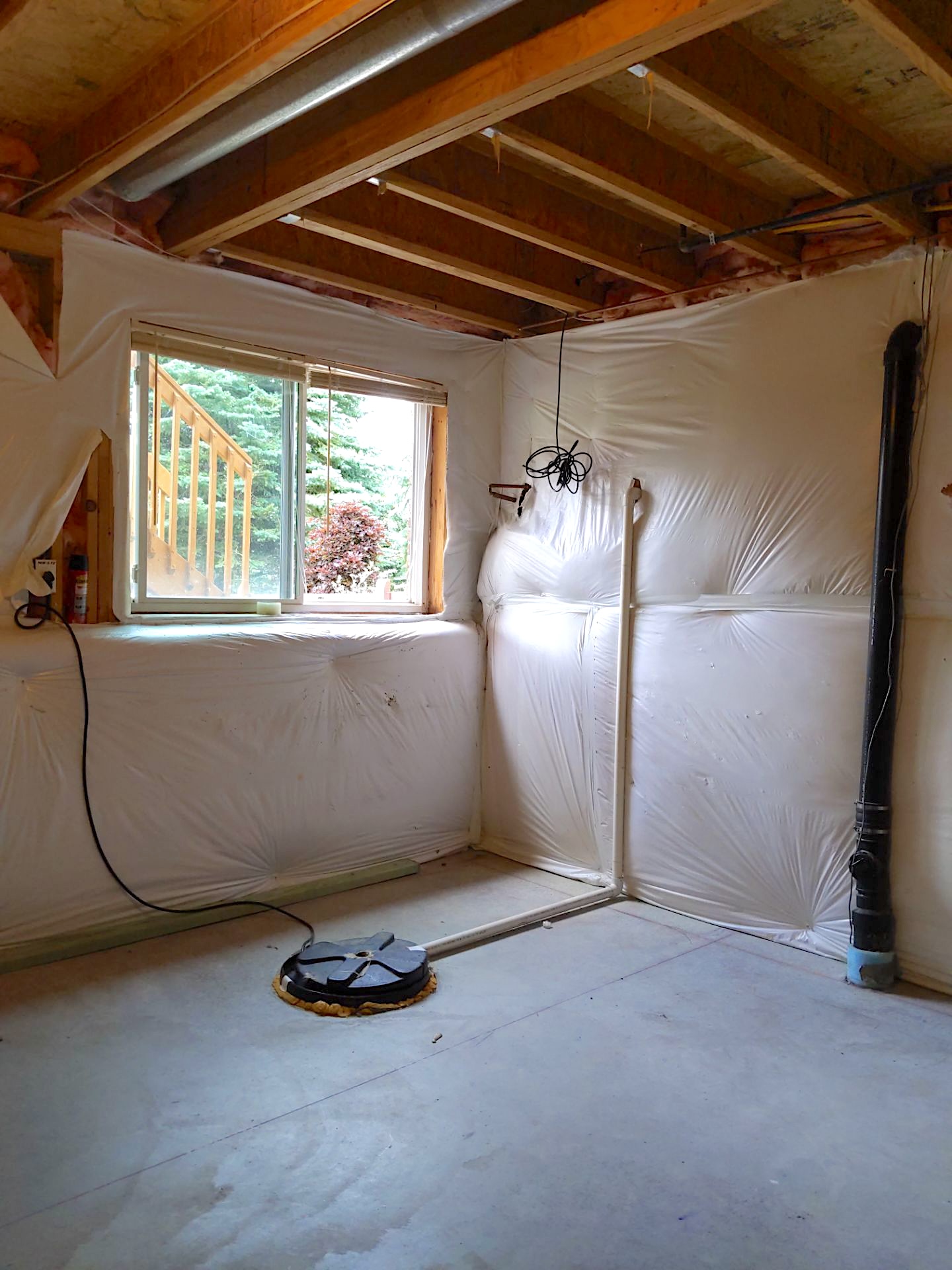 Unfinished basement corner with sump pump, exposed ceiling joists, and plastic wall covering