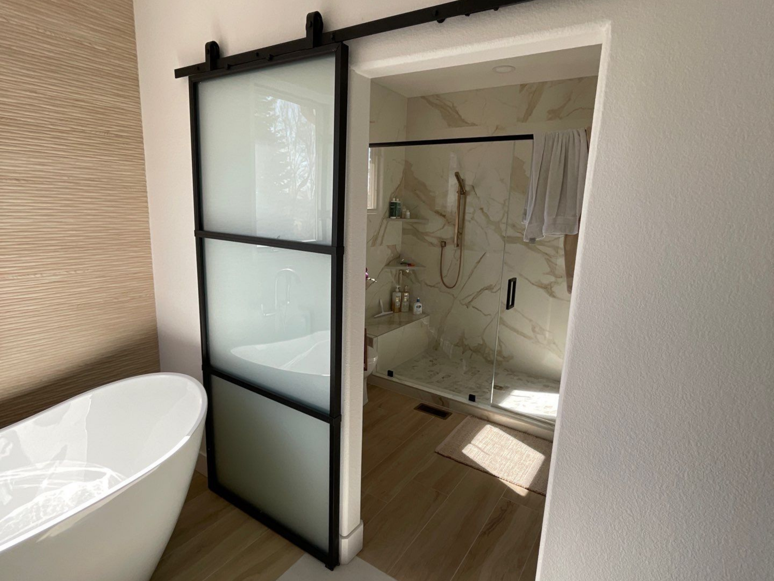 Spa-like bathroom with freestanding tub, modern black-framed sliding barn door, and marble-style walk-in shower with brass fixtures.